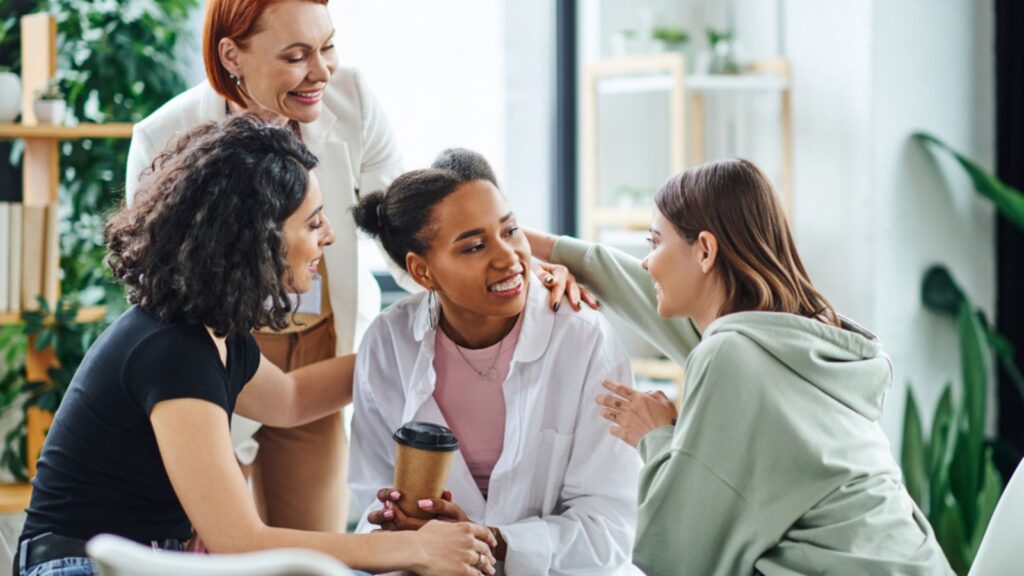 Multicultural women embracing happy african american girlfriend near pleased redhead psychologist during group therapy in consulting room, friendship and mental wellness concept