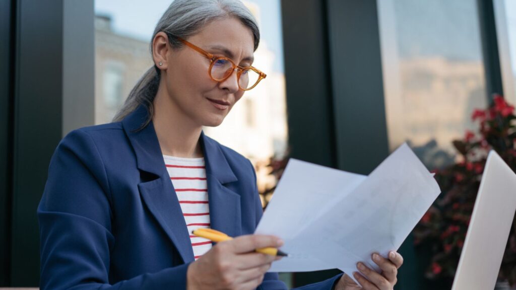 Pensive mature businesswoman reading contract, planning project, brainstorming. Portrait of middle aged asian secretary reading, working with documents, sitting at workplace