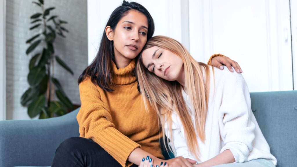 Pretty young woman supporting and comforting her sad friend while sitting on the sofa at home.