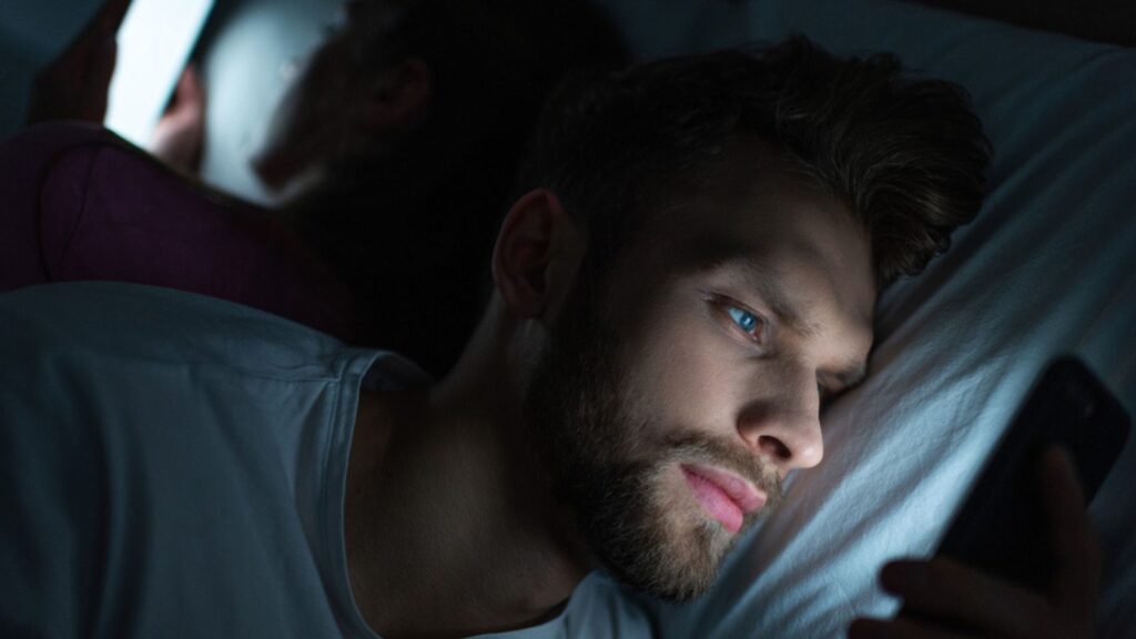 Selective focus of handsome man using smartphone near girlfriend on bed at night