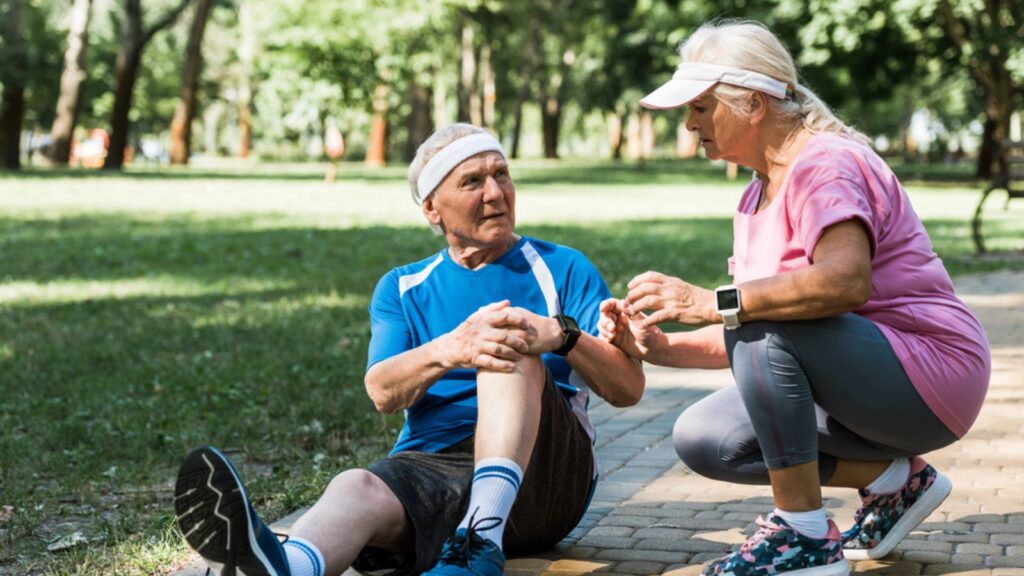 Senior man touching knee while sitting on walkway near retired woman in cap