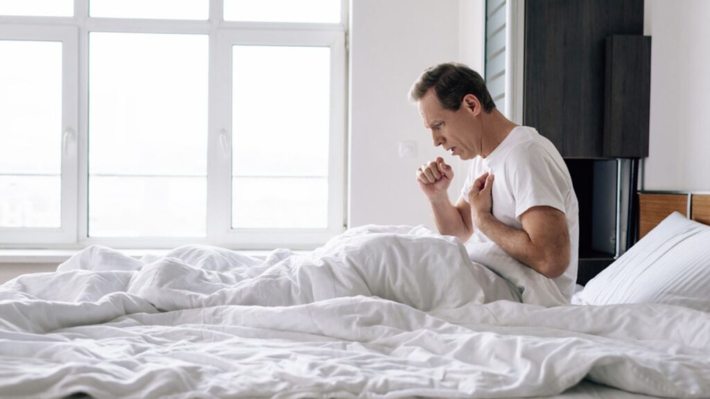 Side view of sick man coughing while sitting on bed at home