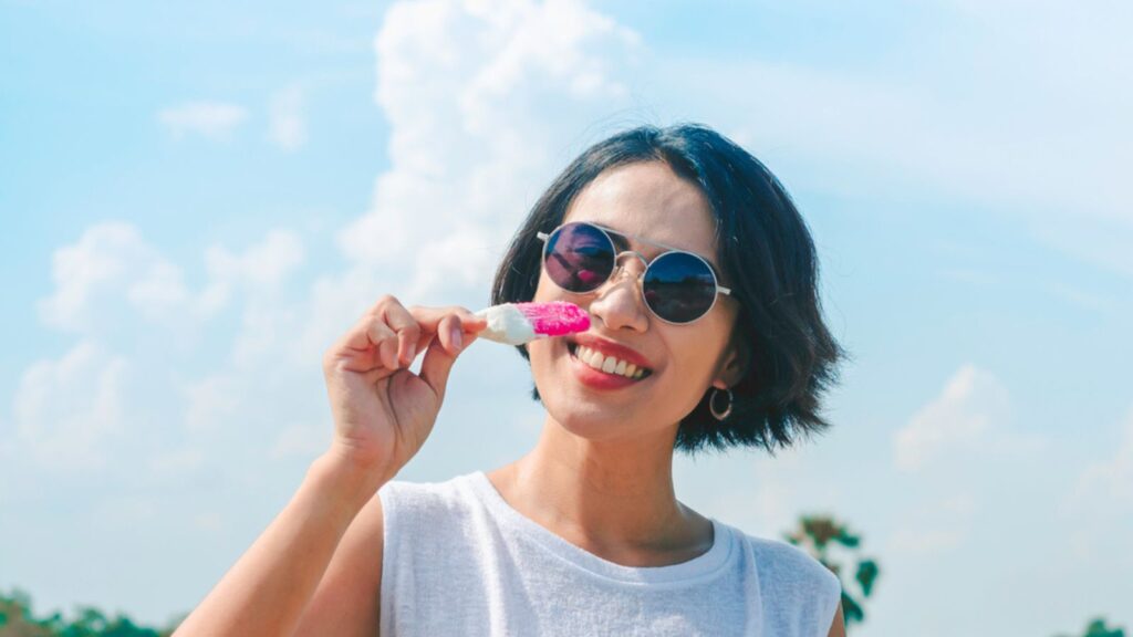 Smiling Asian woman short hair in casual white sleeveless shirt wearing sunglasses holding pink popsicle on blue sky background in summertime.