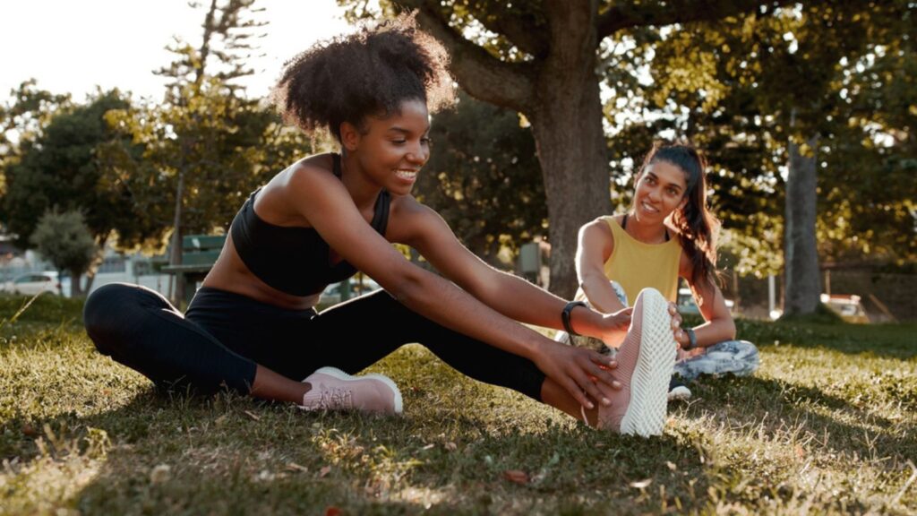 Smiling two diverse sporty fit young female friends stretching their legs in the park