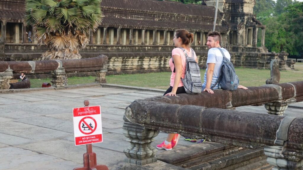 ANGKOR WAT, CAMBODIA, Two disrespectful Caucasian tourists sit on the stone railing next to sign prohibiting people from touching and sitting on the decaying remnants of ancient Angkor