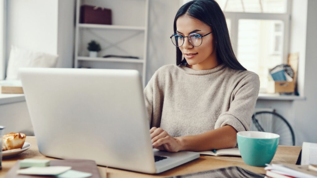 Beautiful young woman in casual clothing using laptop and smiling while working indoors