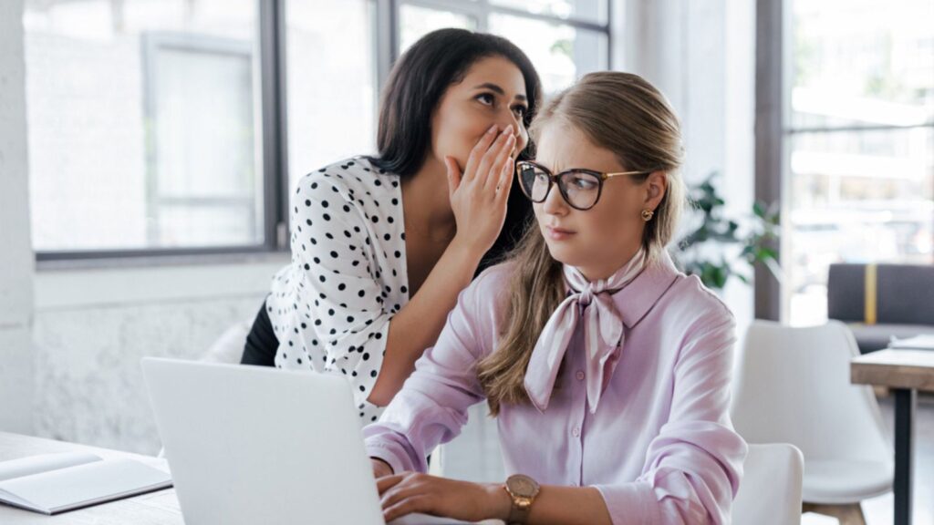 Businesswoman whispering in ear of coworker in glasses while gossiping in office