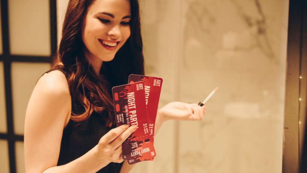 Happy girl in black dress holding lip gloss and night party tickets in bathroom, end of quarantine concept