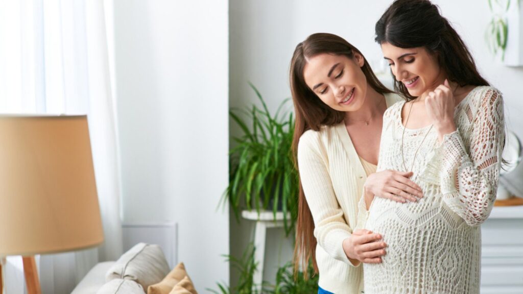 Joyous lgbt couple hugging smiling and looking at pregnant belly, in vitro fertilisation concept