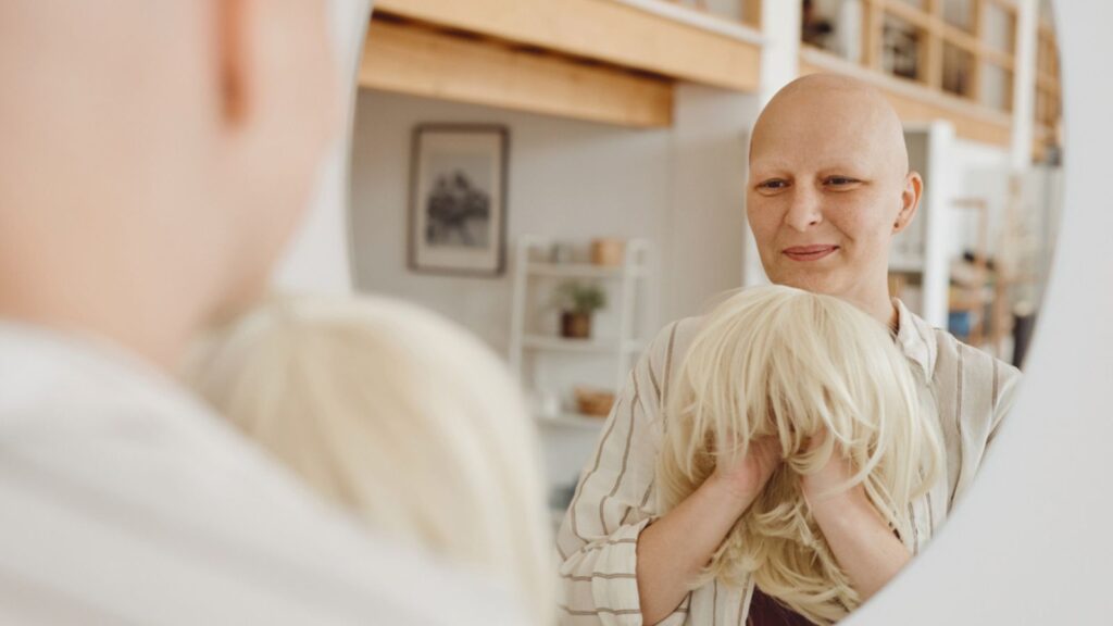 Reflection portrait of bald adult woman looking in mirror holding wig while standing in warm-toned home interior, alopecia and cancer awareness, copy space