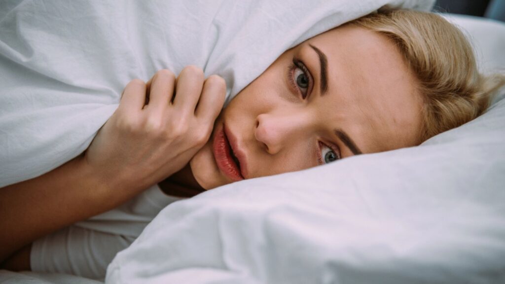 Scared woman covered in blanket looking at camera in bed at home