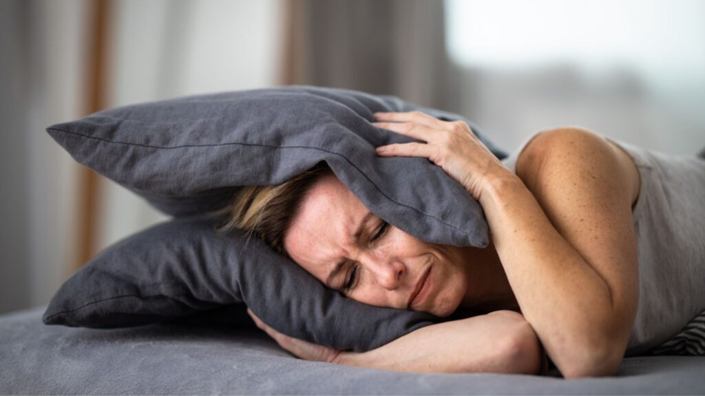 Sleepless lady covering ears with pillow. Noisy neighbors, tinnitus, insomnia or stress concept. Tired woman can't sleep. Awake in bed after coming home from her shift, work