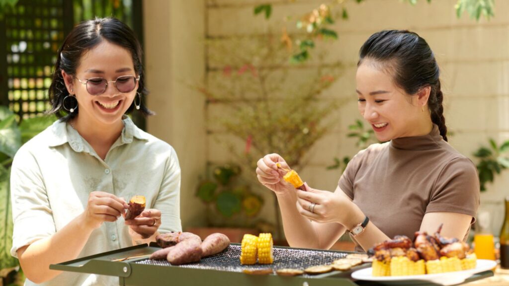 Smiling friends cooking sweet potato and corn on grill