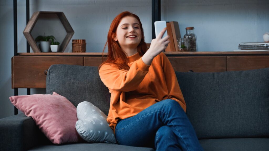 Smiling girl using smartphone near pillows on couch