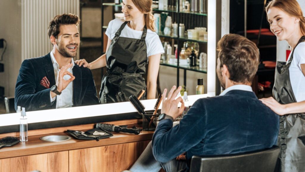 Smiling young hairdresser looking at handsome happy client showing ok sign and looking at mirror in beauty salon