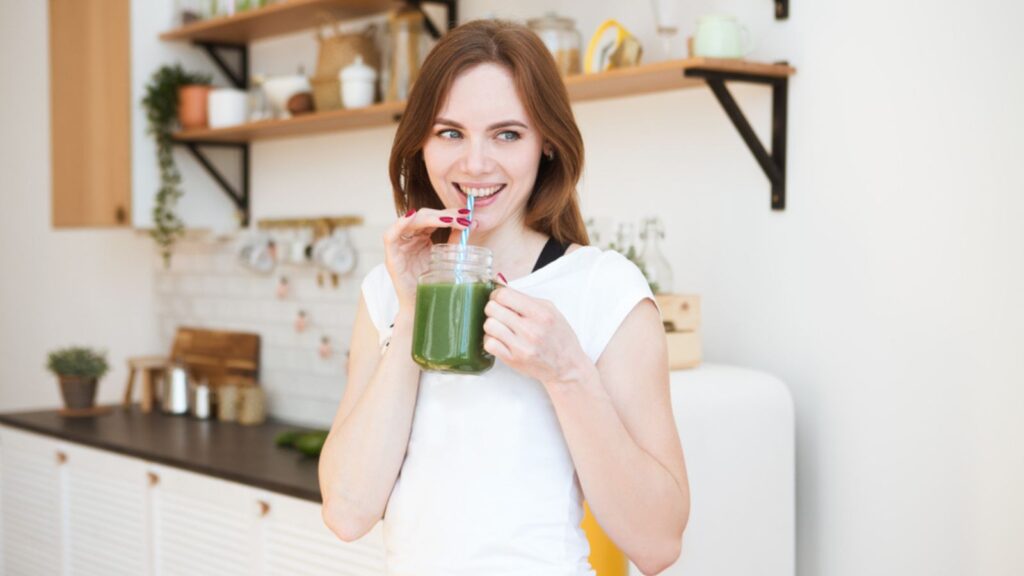 Smiling young woman drinking green smoothie juice in kitchen
