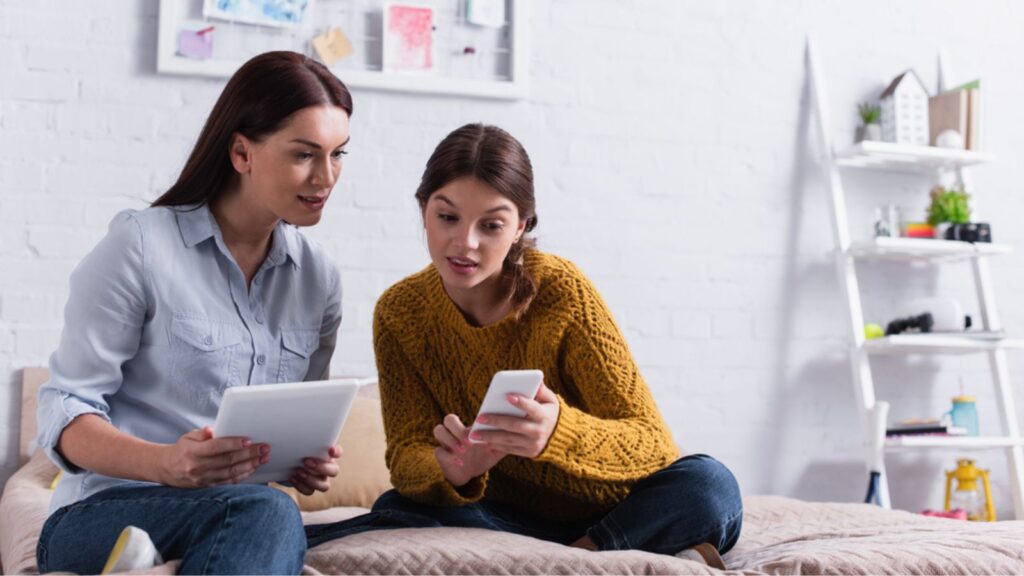 Teenage girl holding smartphone near mother with digital tablet on bed, Teenage girl teaching mother how to use gadget