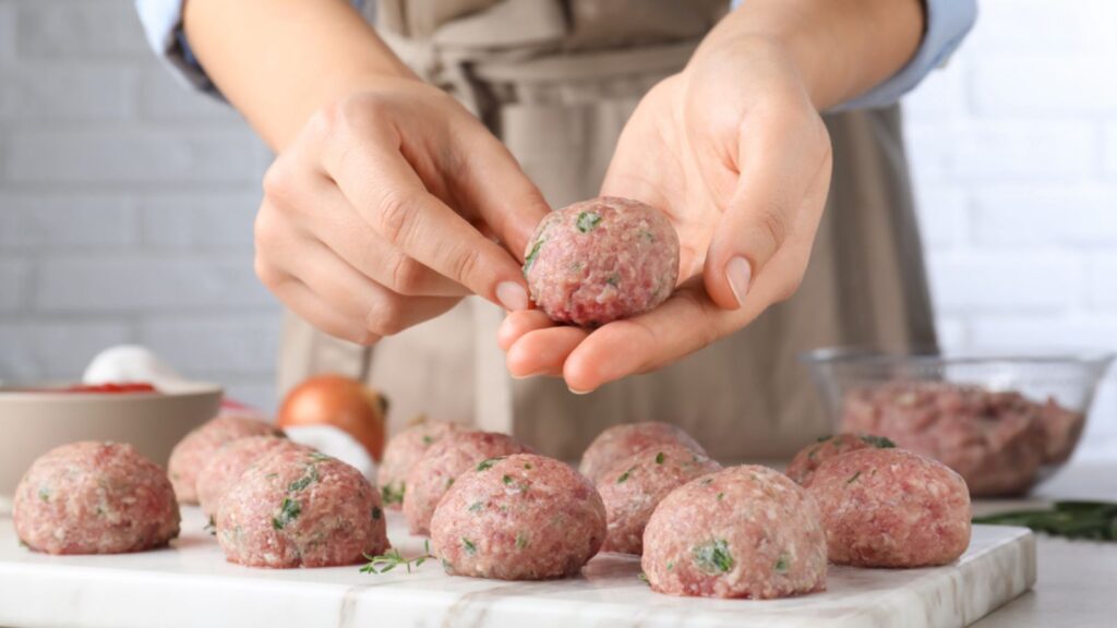 Woman making fresh raw meatballs at white table indoors, closeup
