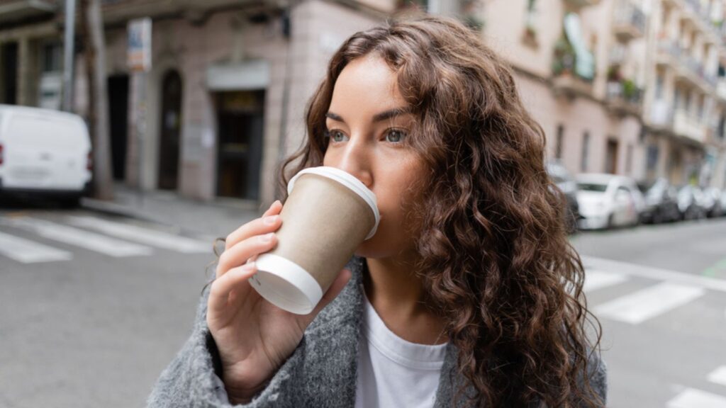woman in casual jacket drinking coffee to go from paper cup and looking away while standing near blurred building on city street in Barcelona, Spain