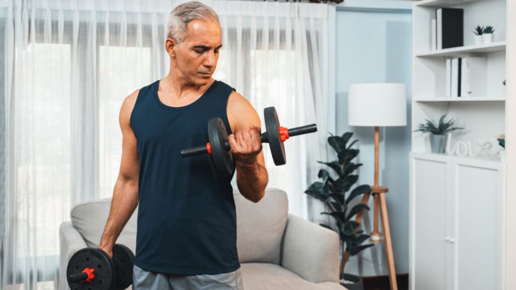 Athletic and sporty senior man engaging in body workout routine with lifting dumbbell at home as concept of healthy fit body with body weight lifestyle after retirement. Bicep curls