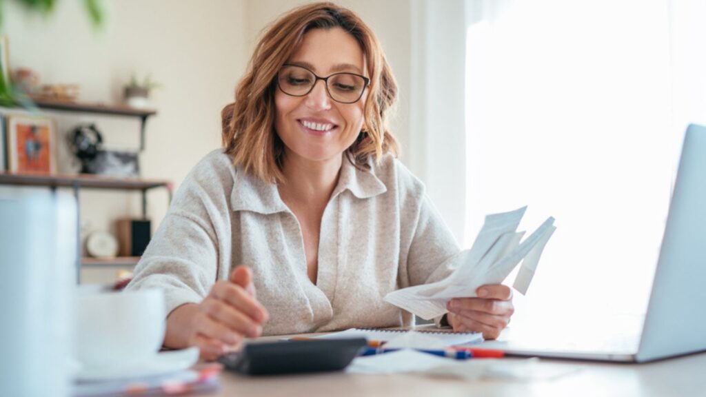 Beautiful middle-aged woman in glasses and paper bills smiling while calculating revenue grows at home office