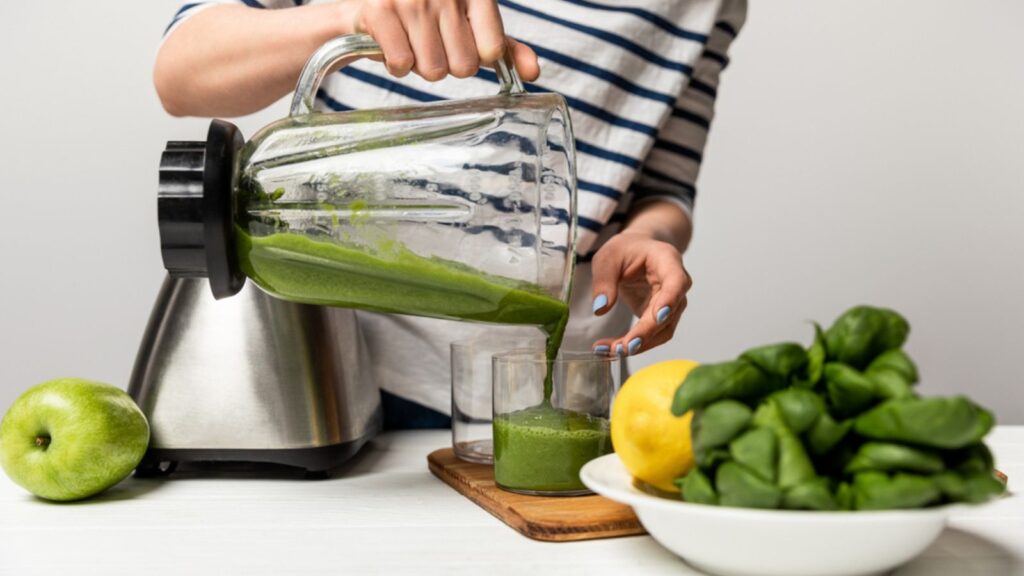 Cropped view of woman pouring green smoothie in glass near apple on white