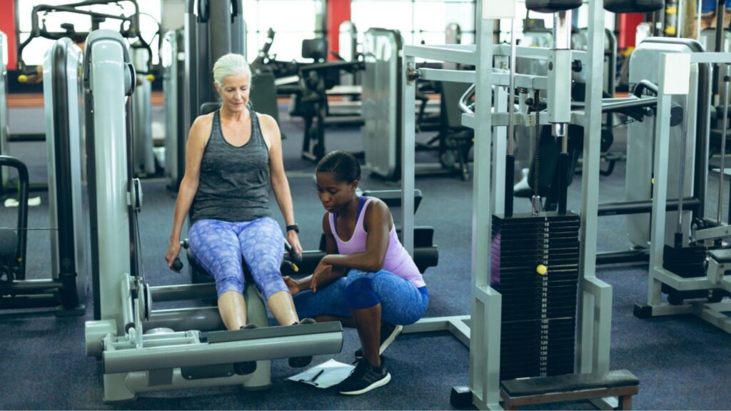 Front view of young pretty African-american female trainer assisting active senior Caucasian woman to work out on leg curl machine in fitness center