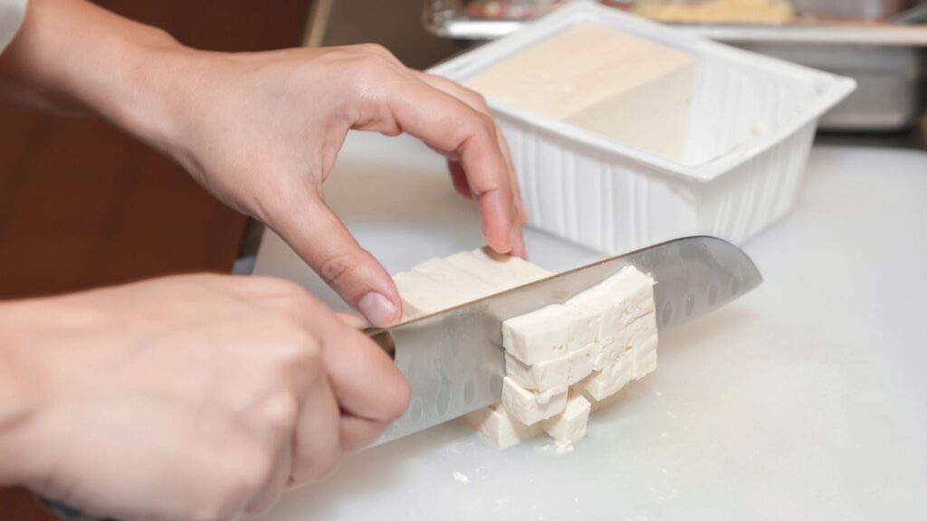 Hands shown cutting a tofu block into pieces with a knife
