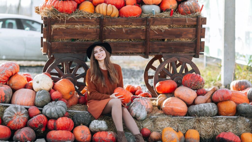 Happy smiling woman sitting with pumpkin in hands near wagon with orange pumpkin on farmers market in brown sweater, dress and hat. Cozy autumn vibes Halloween, Thanksgiving