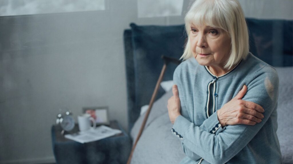 Lonely senior woman sitting on bed with hands crossed at home