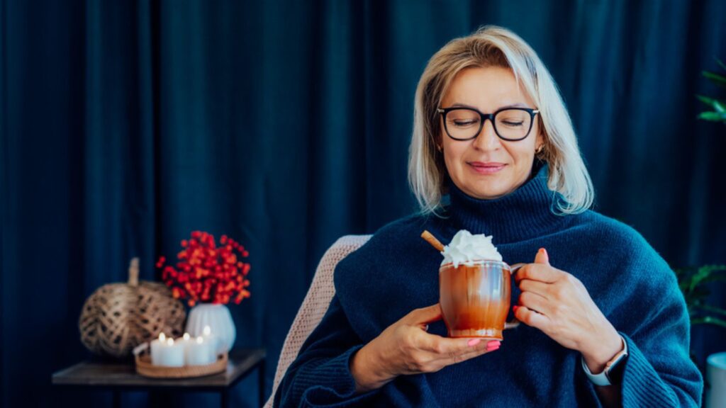 Middle aged woman relaxing with cup of hot drink in scandy style hygge interior home with fall mood interior decor. Lady dreaming, enjoy calm mood without stress, wellbeing alone.