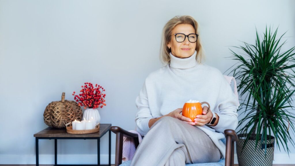 Middle aged woman relaxing with pumpkin shaped cup of hot drink in scandy style hygge interior home with fall mood decor. Lady dreaming, enjoy calm mood without stress, well being alone.