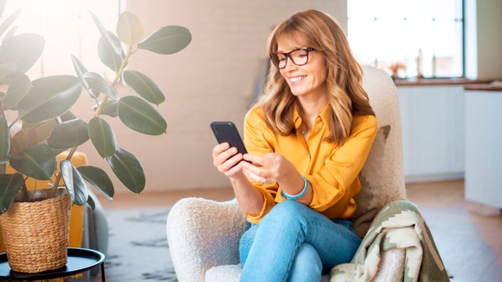 Portrait shot of happy middle aged woman using mobile phone and text messaging while relaxing in the armchair at home