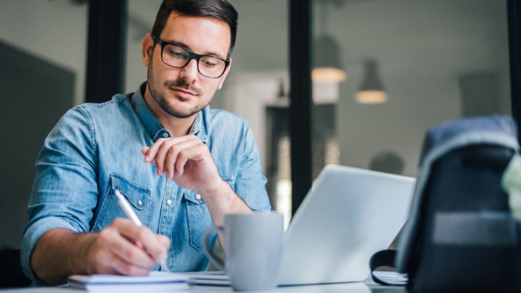 Close up portrait of handsome man working from home office taking reading and writing notes in note pad while working on laptop computer