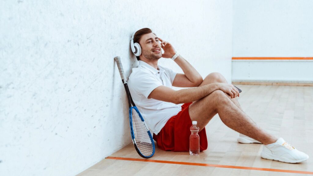 Smiling squash player sitting on floor and listening music in headphones