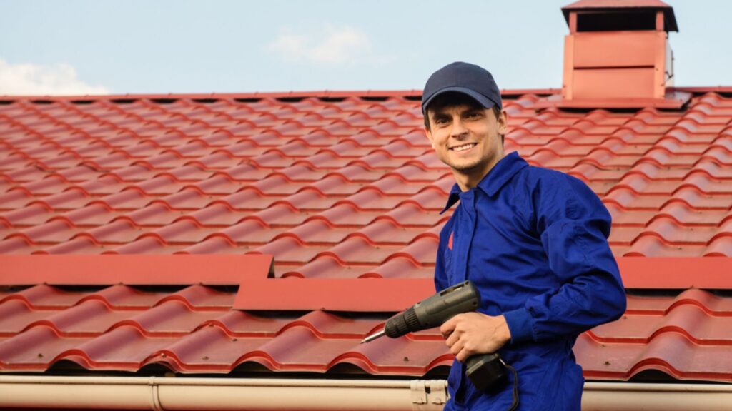 Young happy man contractor worker in blue overalls is repairing a red roof with electric screw driver