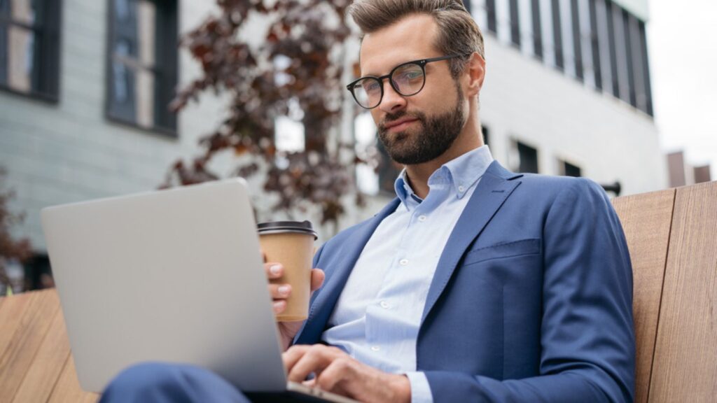 man wearing suit, stylish eyeglasses holding cup of coffee watching training courses. Confident businessman using laptop computer working online