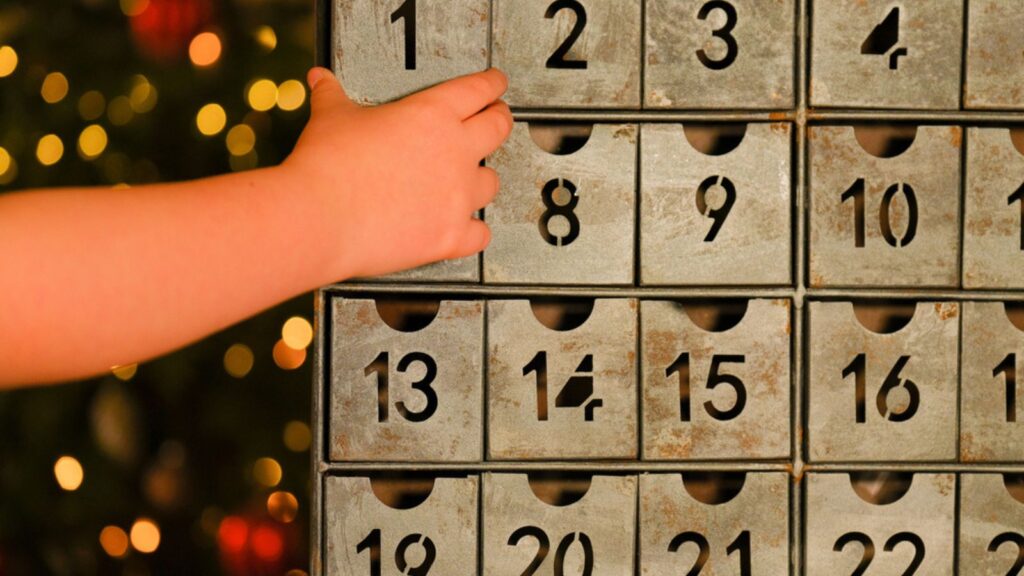 Advent calendar in vintage style and childs hand near Christmas tree with toys glowing garland on blurred room background.Christmas traditions and symbols