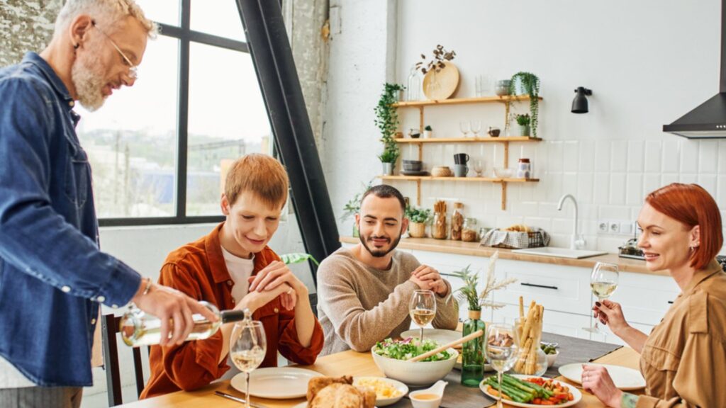 Bearded man pouring wine near delicious supper and happy family in modern kitchen