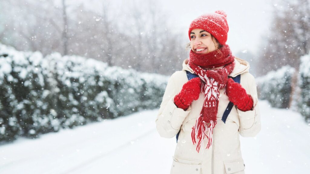 Cheerful smiling woman in warm clothes, red knitted cap, scarf and mittens walking on the snowy street under falling snowflakes after blizzard in city. Happy woman playing with the snow