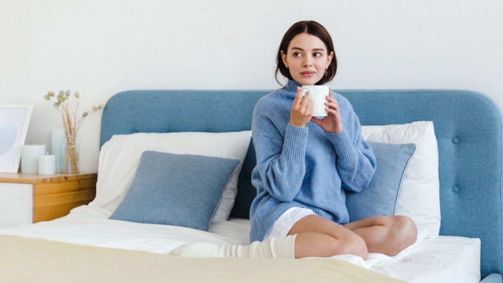 Girl in a blue sweater in interior Hygge style with a cup of hot tea in her hands sits on the bed