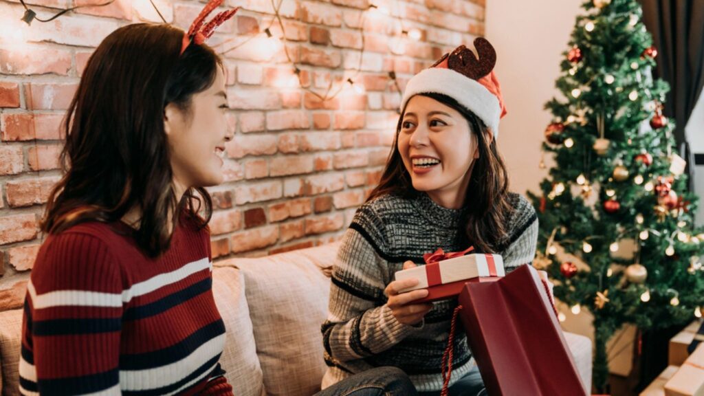 Girl with santa hat got a gift from another girl in red sweater. young best friends exchanging christmas gifts at home. colorful xmas tree standing in the background.