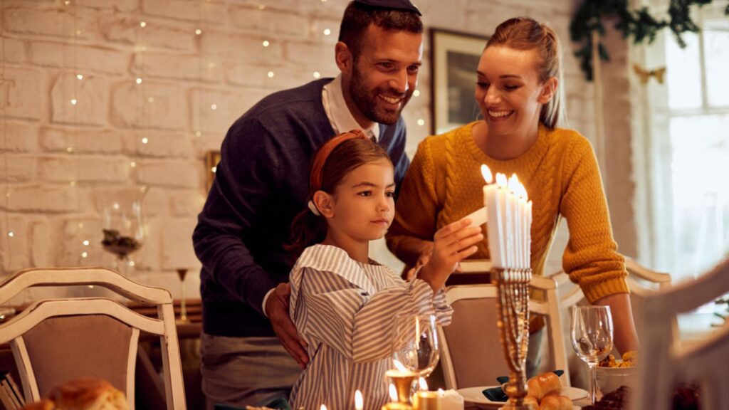 Happy parents with daughter lightning candles in menorah at dining table while celebrating Hanukkah at home.