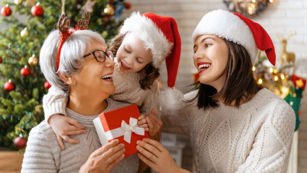 Merry Christmas and Happy Holidays! Cheerful kid presenting gifts to mom and granny. Parents and little child having fun near tree indoors. Loving family with presents in room.