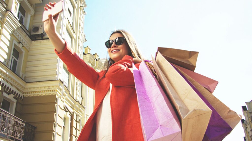 Selfie time. Portrait of cheerful and attractive woman in red coat and sunglasses taking a selfie with shopping bags