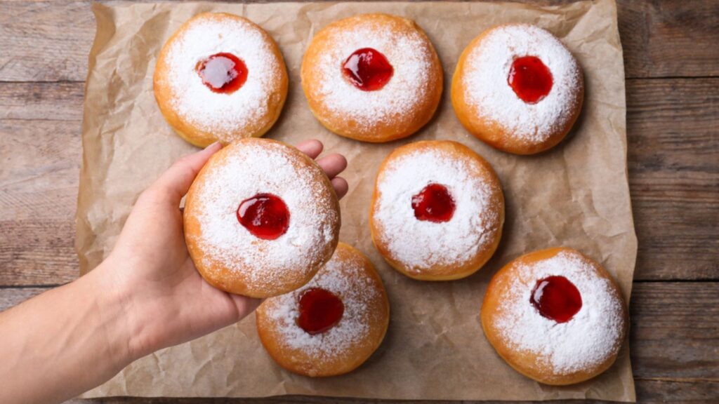 Woman holding Hanukkah doughnut with jelly and sugar powder over wooden table, top view.