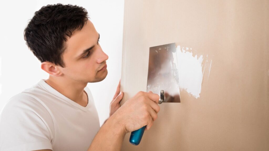 Young man using putty knife on white wall at home