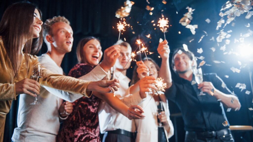 Having fun with sparklers. Confetti is in the air. Group of cheerful friends celebrating new year indoors with drinks in hands